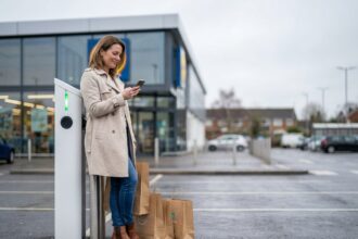 Femme utilisant une borne de recharge électrique dans le parking d'un centre commercial tout en consultant son téléphone